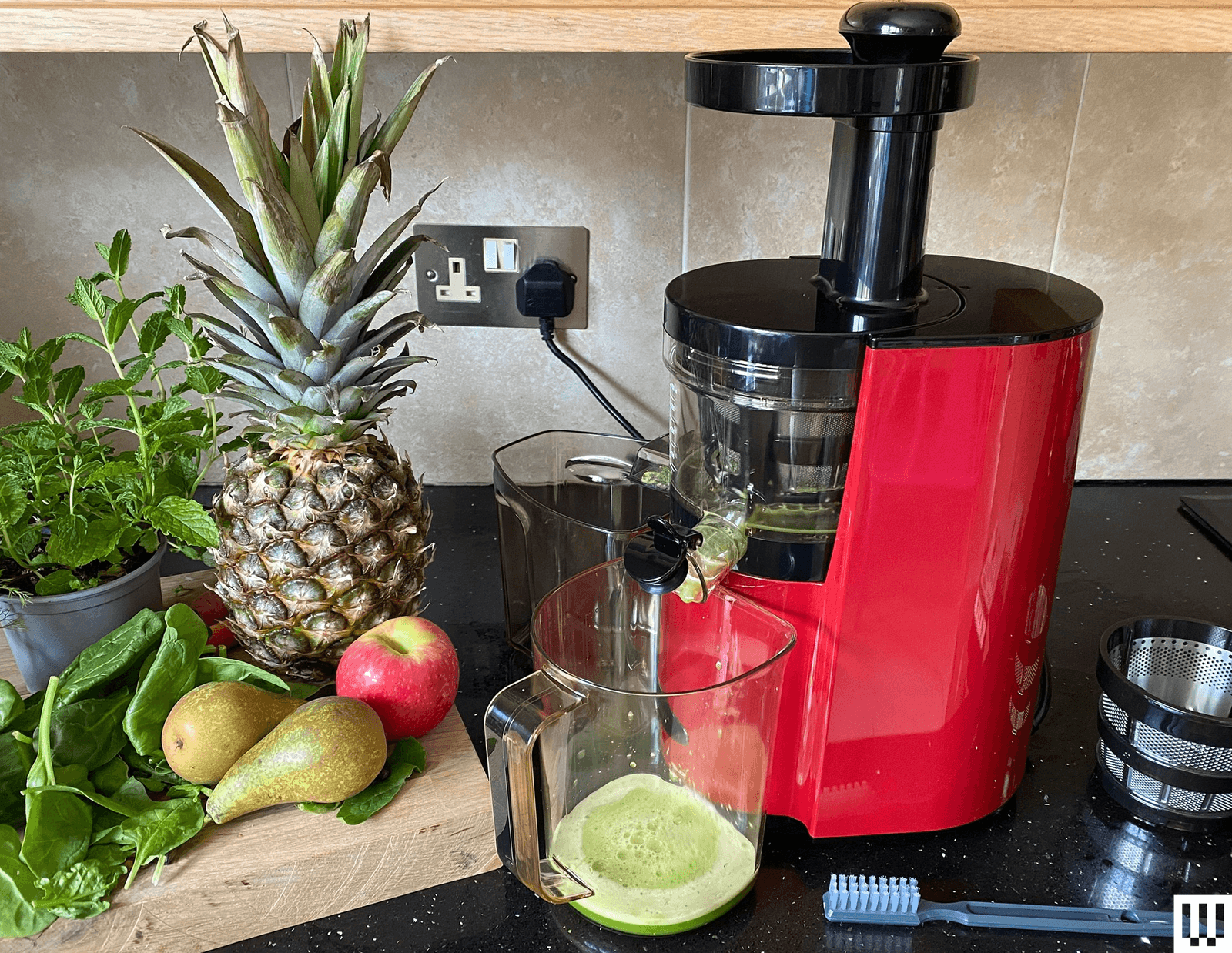 Kitchen table has a cutting board on the left for fruits and vegetables and a red cylindrical juicing machine on...