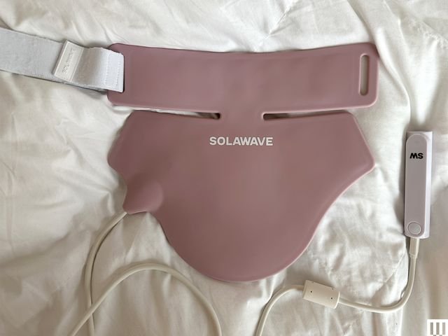Solawave Neck and Chest Rejuvenating Mask, a pink latex face mask that lies flat with a wire attached on top of a white blanket