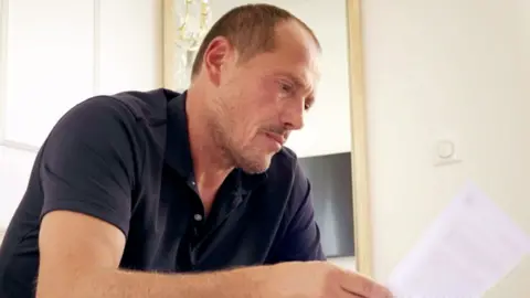 BBC A man with short, light brown hair and stubble looks at a piece of paper. He is wearing a dark blue, short-sleeved polo shirt. 