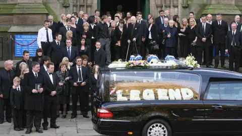 Getty Images Funeral mourners stand on the church steps while a hearse sits outside. A floral greeting reads the word 