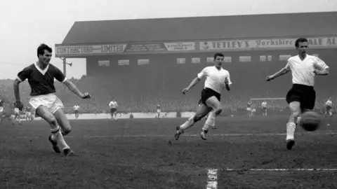 PA Media Dougan watches his shot go towards goal at a match in front of a crowd. Two opposing players wearing white run towards the ball. The pitch is very muddy.