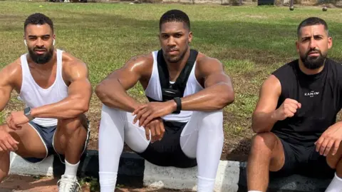 Social Media Latif Ayodele sits with Anthony Joshua and Senna Gami on the grass outside, all wearing training gear