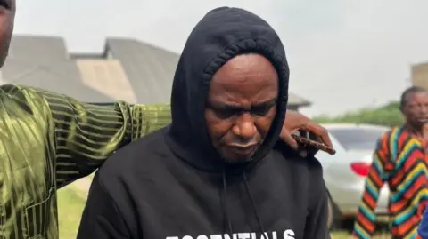 BBC Adeniyi Kayode wears a black hoodie and looks down. Another man, further out of the picture, has his arm around Mr Kayode