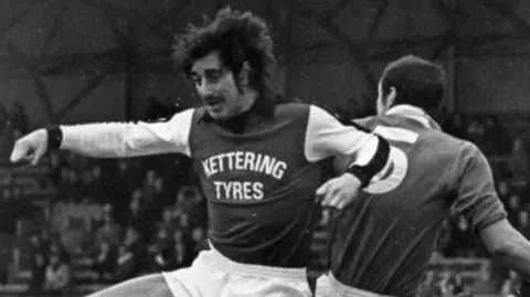 Kettering Town FC Black and white photo of a Kettering Town footballer in action. He competes in the air for the ball that is out of shooting range. An opposing player stands behind him, as does the crowd.