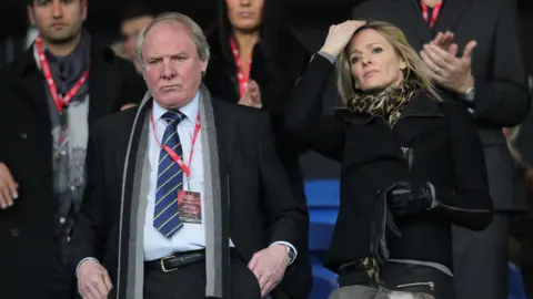 PA Media A man and a woman stood side by side looking forward to a soccer match, a look of concentration on both faces. The man wears a blue shirt and tie, while the woman wears a black coat and leopard-print scarf. 