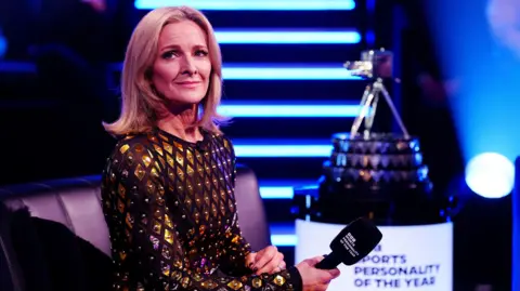 PA Media Gabby Logan wears a black dress with a gold sequence spread across it. She holds a microphone in her right hand and the Sports Personality of the Year award can be seen blurred in the background to her right.