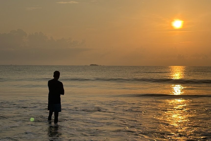 A person standing in water at a beach at sunset.