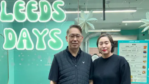 Hyung Joon Kim A middle-aged couple stands outside the glass-fronted restaurant named after... 