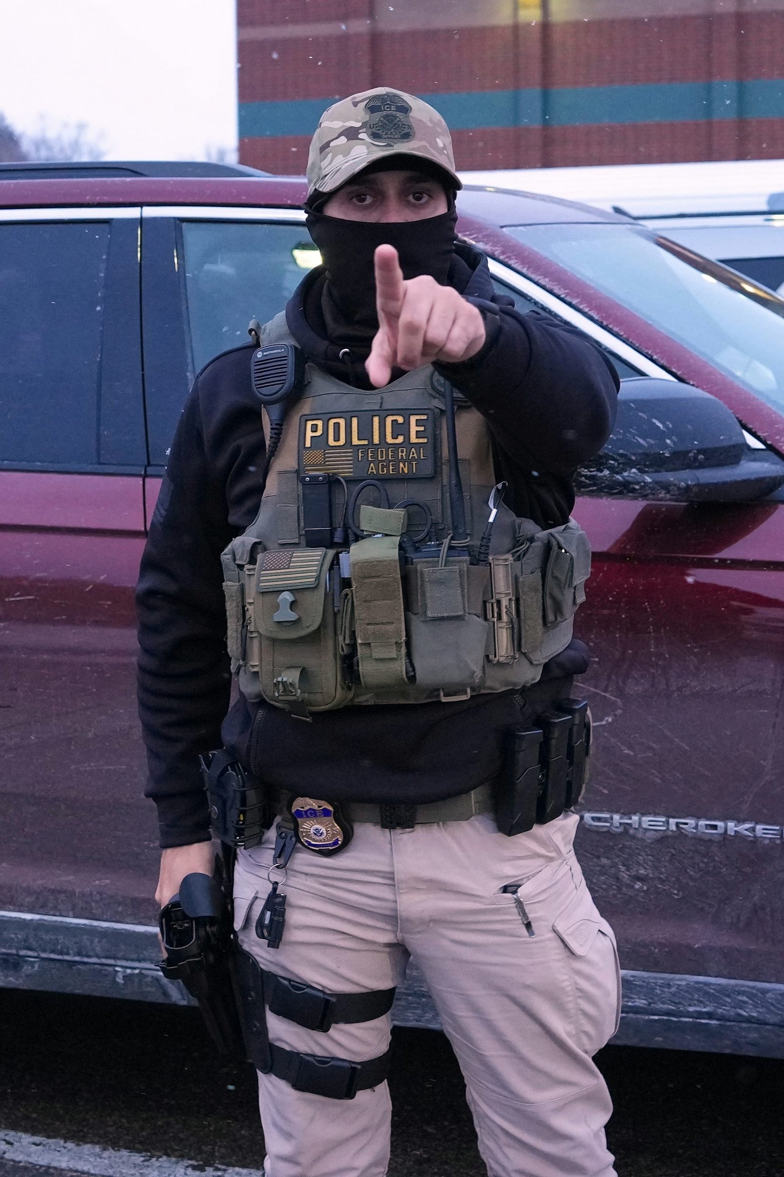 a man wearing a tactical vest and face mask points in the direction of the viewer