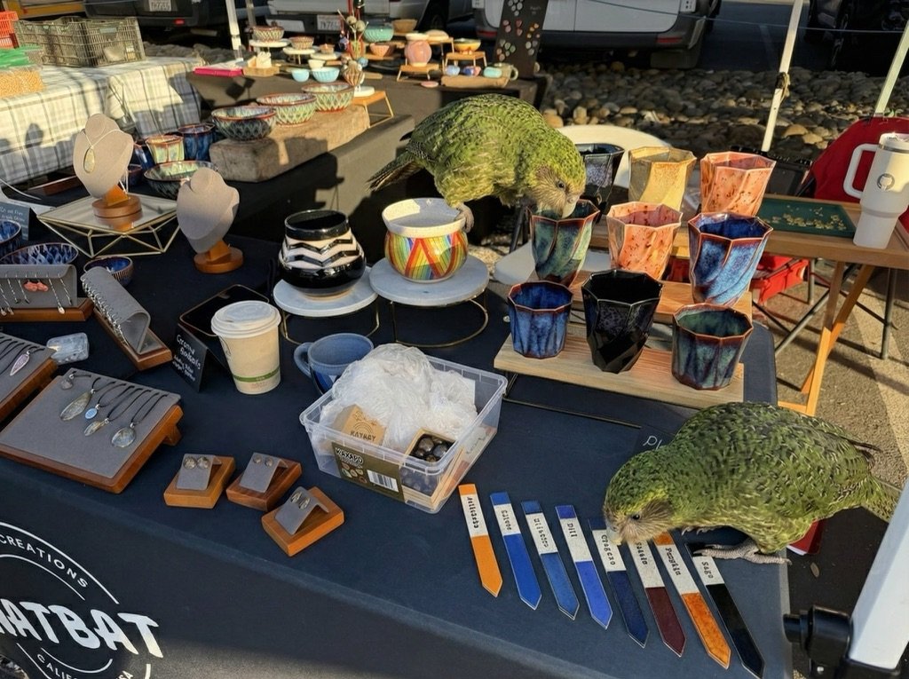 Craft market booth with ceramics and two kākāpō. One is center-table peering into ceramic cups near a rainbow pot, while the second is at the right edge of the table near the plant markers, appearing to examine or possibly chew on items at the table