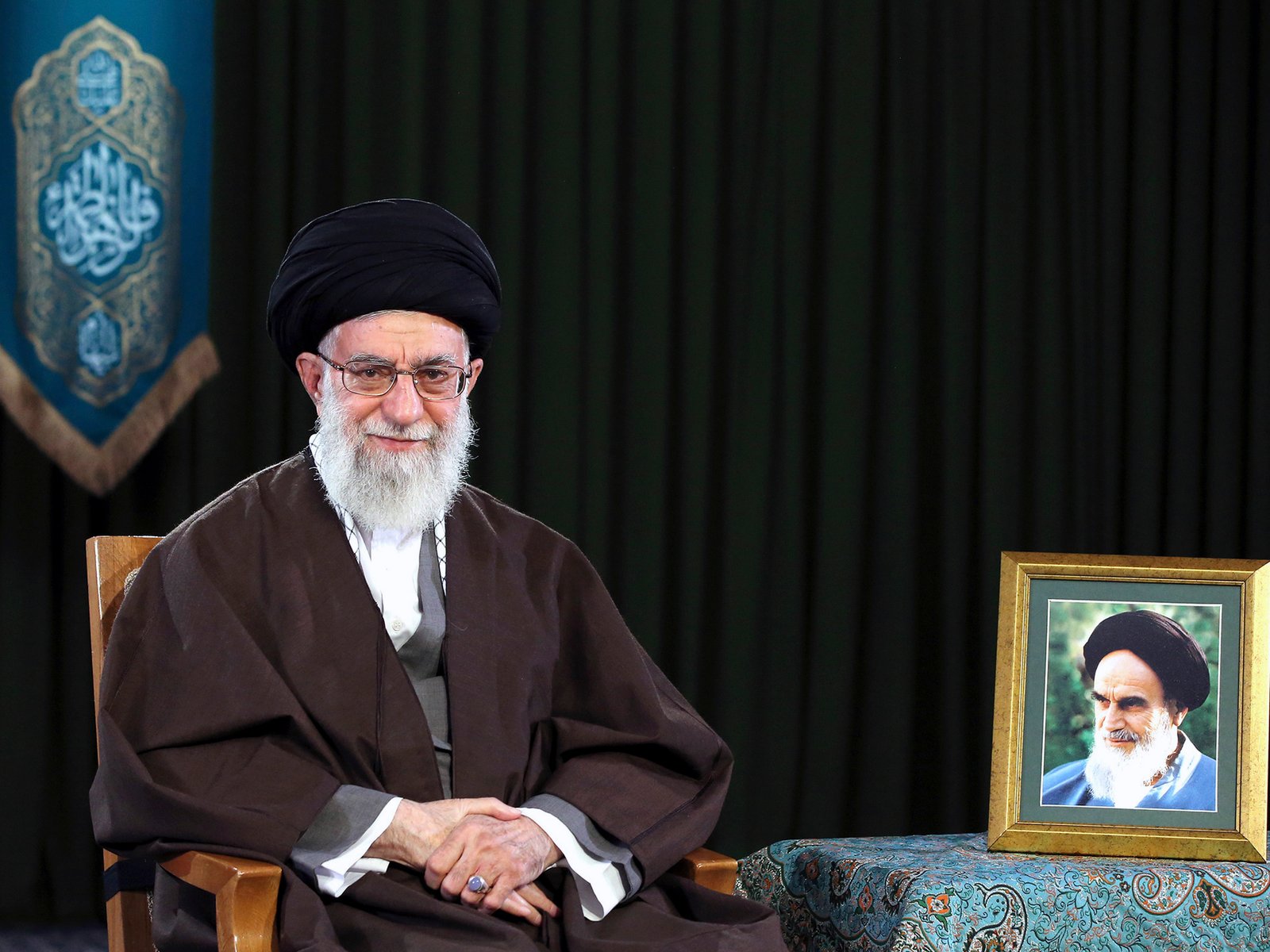 In this 2017 photo, Ayatollah Ali Khamenei, wearing a turban and traditional clothing, sits on a chair. A framed portrait of Ayatollah Ruhollah Khomeini stands on a table on the right side of the frame.