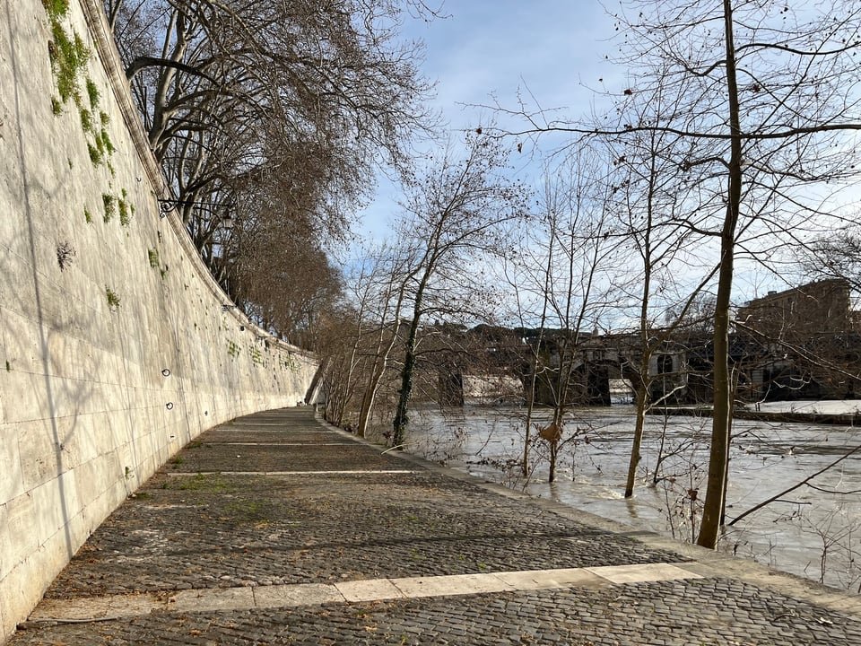 A tall embankment wall rises on the left, a river on the right, and a cobblestone road in the middle. Trees are partially submerged on the edge of the river. In the distance, Roman bridge ruins.