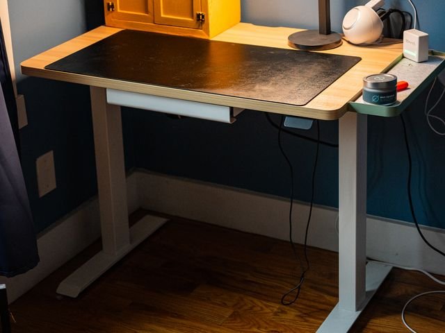 A Branch Duo standing desk with a tan top and white legs includes a small cabinet, a black cutting mat and a lamp on top