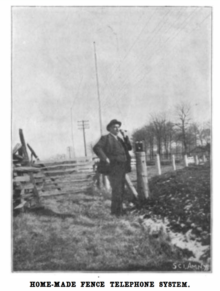 black and white photograph of a pan in a suit and bowler hat standing in front of a barbed wire fence talking on the phone