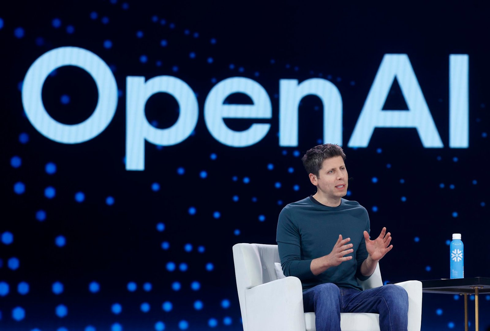 A man speaks while sitting in a chair in front of a wall emblazoned with OpenAI.
