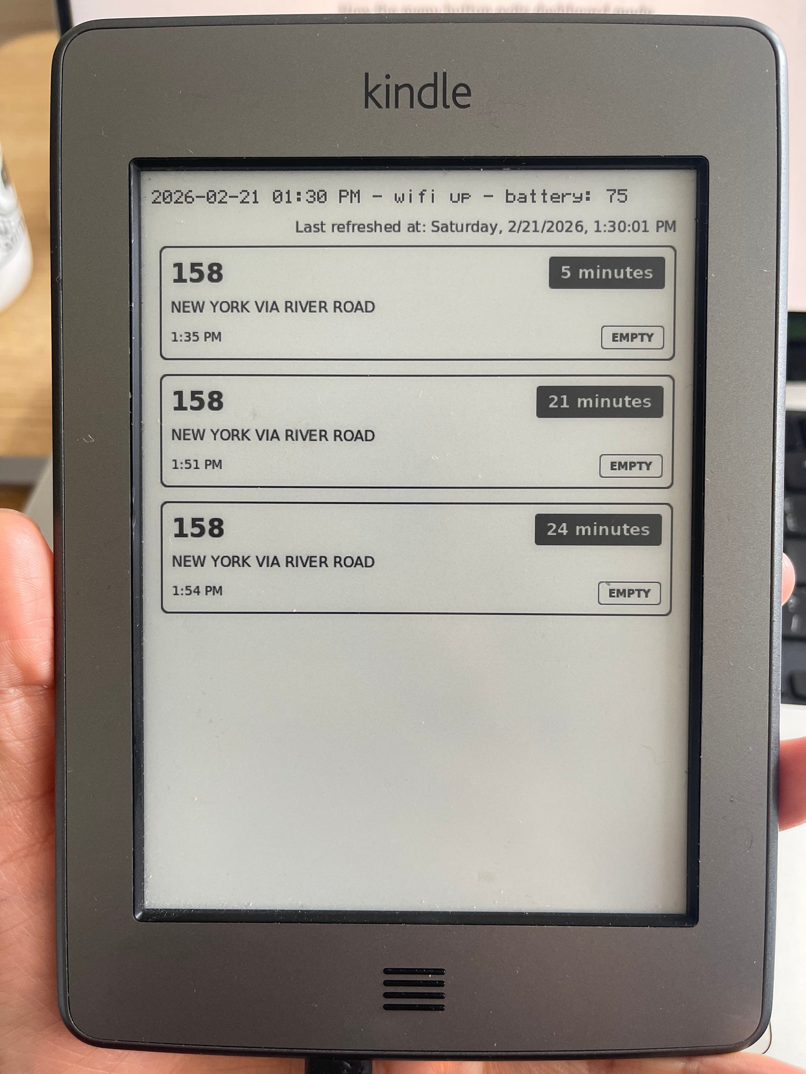 A closeup of the Kindle screen, with a list of 3 upcoming busses, along with current date, wifi status, and battery amount displayed up top