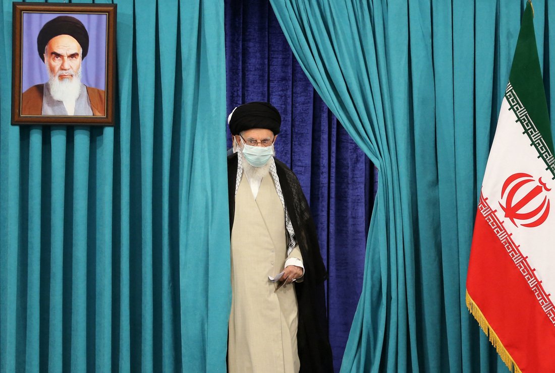 Emerging through a pair of teal curtains, Ali Khamenei wears a mask due to the COVID-19 pandemic as he arrives to cast his ballot during Iran