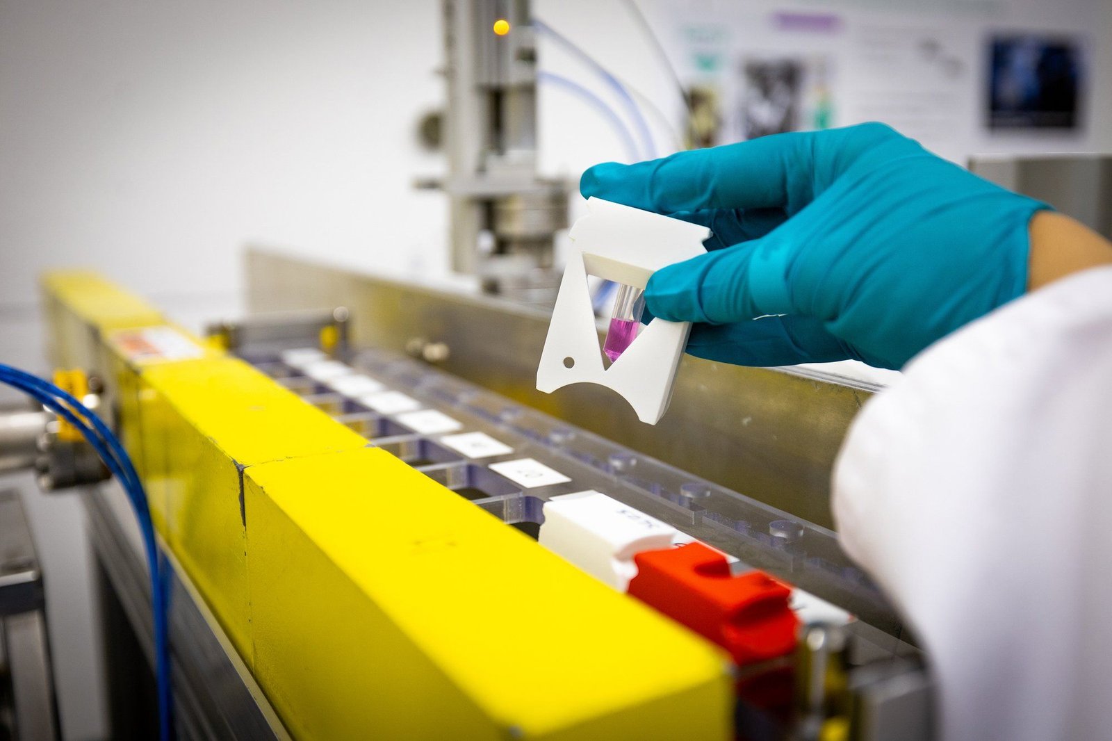A photo of a closeup of a gloved hand holding a sample of a purple liquid above a piece of equipment.