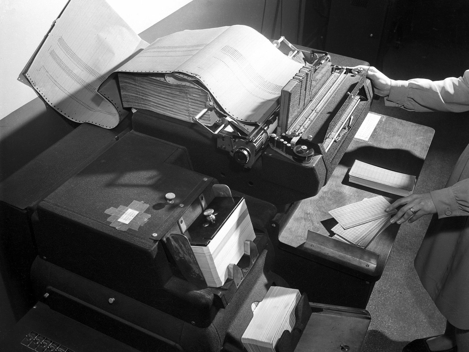 A white woman using a computer-adding machine in the 1940\u2019s. The device resembles a bulky typewriter and prints large stacks of paper with tabulated answers.