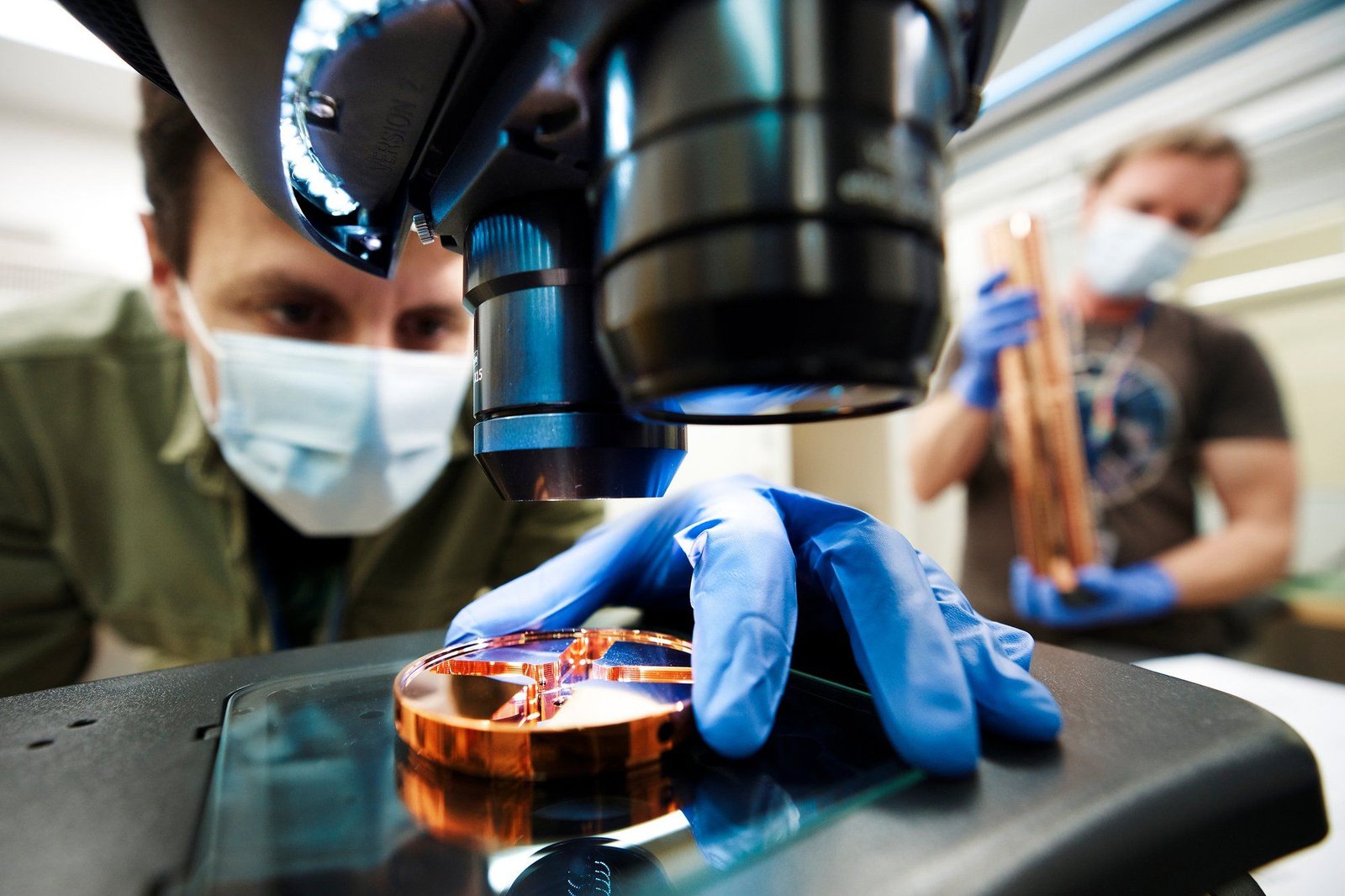 Close-up photo of an etched copper disc being held under a microscope by a gloved hand.