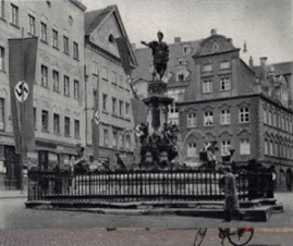 Adolf-Hitler-Platz in Augsburg with Nazi parade archival footage. Reichsadler stone eagle on Augsburg Landratsamt building facade. Unrealised Nazi Gauforum architectural plans by Hermann Giesler. Messerschmitt Bf 109 aircraft factory ruins after 1944 bombing. Augsburg synagogue building now housing Jewish Cultural Museum. Hall 116 memorial at former Augsburg subcamp site.
