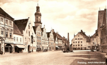 Adolf-Hitler-Straße, Freising's main street (now Obere Hauptstrasse). Marienplatz square in Freising where Nazi rallies were held under swastika banners. Former Adolf-Hitler-Straße main thoroughfare in Freising during Third Reich. Freising Cathedral on Domberg showing complex Catholic Church role under Nazism. Site of former Lewin family department store targeted in Nazi boycotts. Freising air raid destruction from 1945 Allied bombing shown in archival photos. Modern Freising street with extremist graffiti and vandalism on buildings.