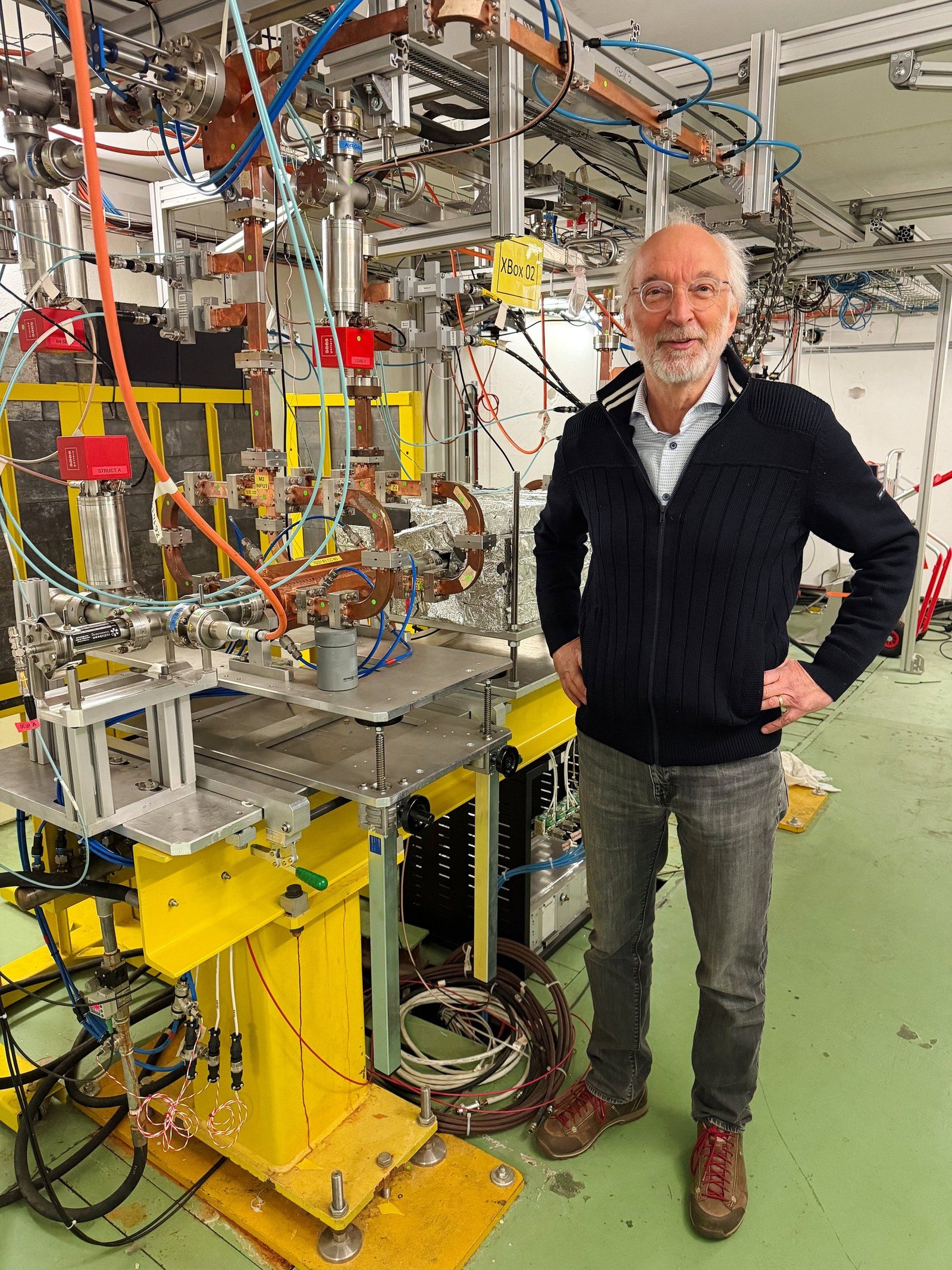 Photo of a white-haired man standing next to floor-to-ceiling experimental equipment with many tubes and wires. 