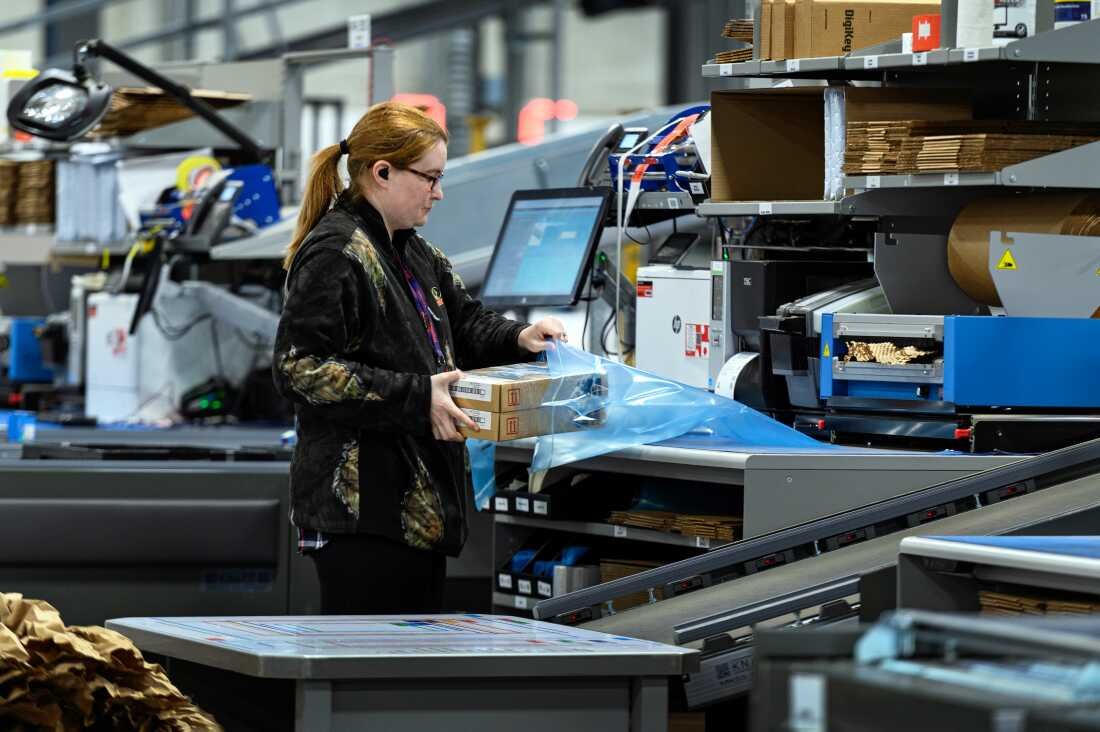 In this photo, a woman at a workstation packs two small cardboard boxes into a large transparent bag.