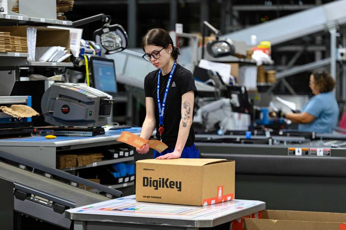In this photo, a young woman wearing a black T-shirt attaches a label to a cardboard box. Near her is a conveyor belt.