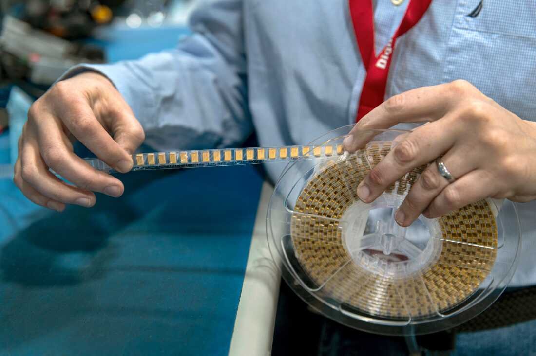This close-up photo shows a reel around which is spooled individual electronic components, each of which is a small gold-colored rectangle attached along a clear ribbon. One hand is holding the reel, and another is unspooling the ribbon.