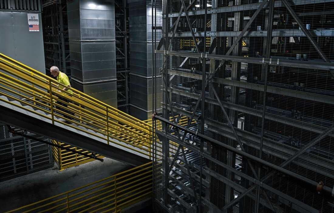 This photo shows the automated storage and retrieval system in DigiKey’s product distribution center, which is four stories high. The space has tall, long rows of metal shelves. A worker walks up a staircase with yellow handrails.